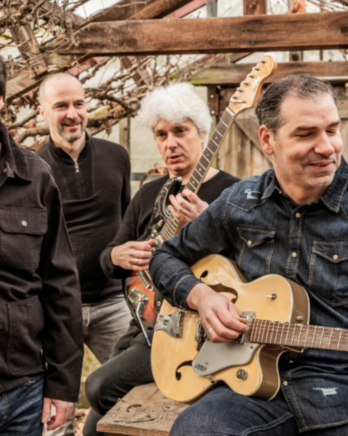 Four middle-aged men sitting outside wearing monochrome clothing, two of them holding an electric guitar in what seems to be a barn