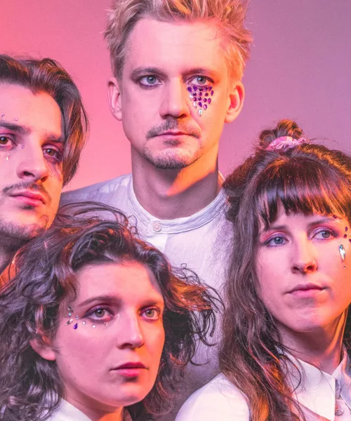 Portrait of two men and two women with crystals on their face looking away from the camera sitting in front of a pink background