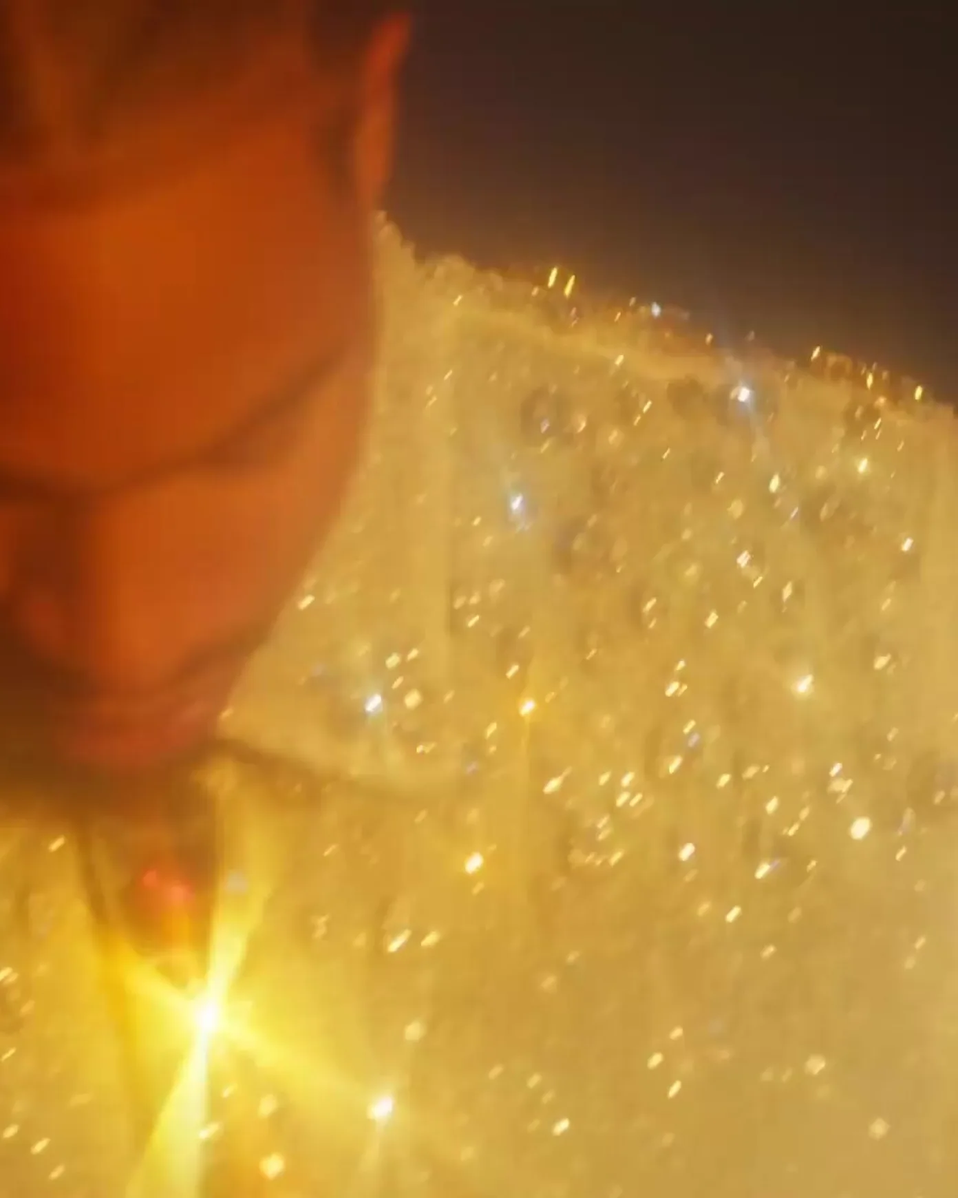 Soft closeup of a braided man with a trimmed facial hair looking downwards, the man is wearing a white shirt covered with crystals and the image has a warm hue to it