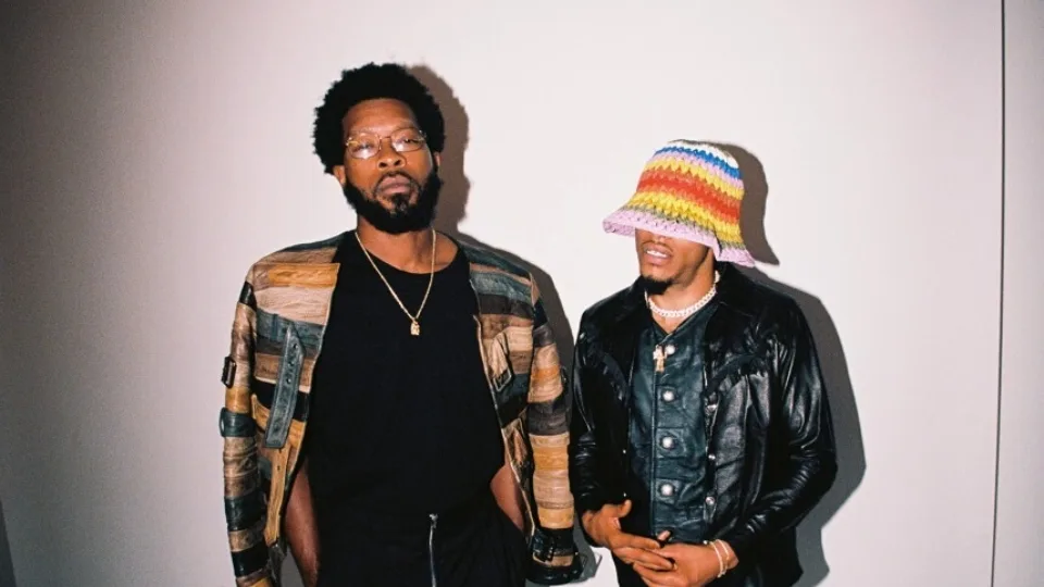 Two men standing in front of a white wall, one with glasses and a colorful jacket looking straight in the camera and the other one looking away with a colorful hat covering his eyes