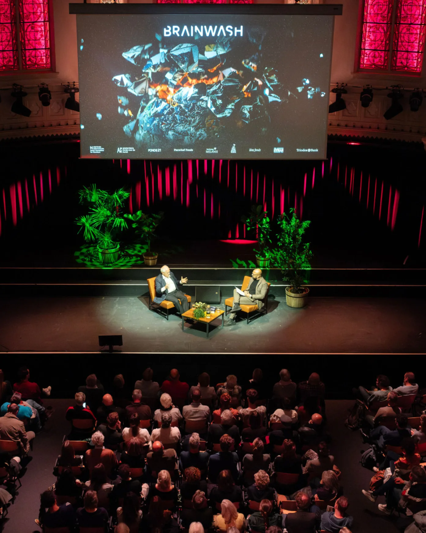 Two men sitting on a podium talking in front of an audience amidst red and green lighting and a big screen behind them