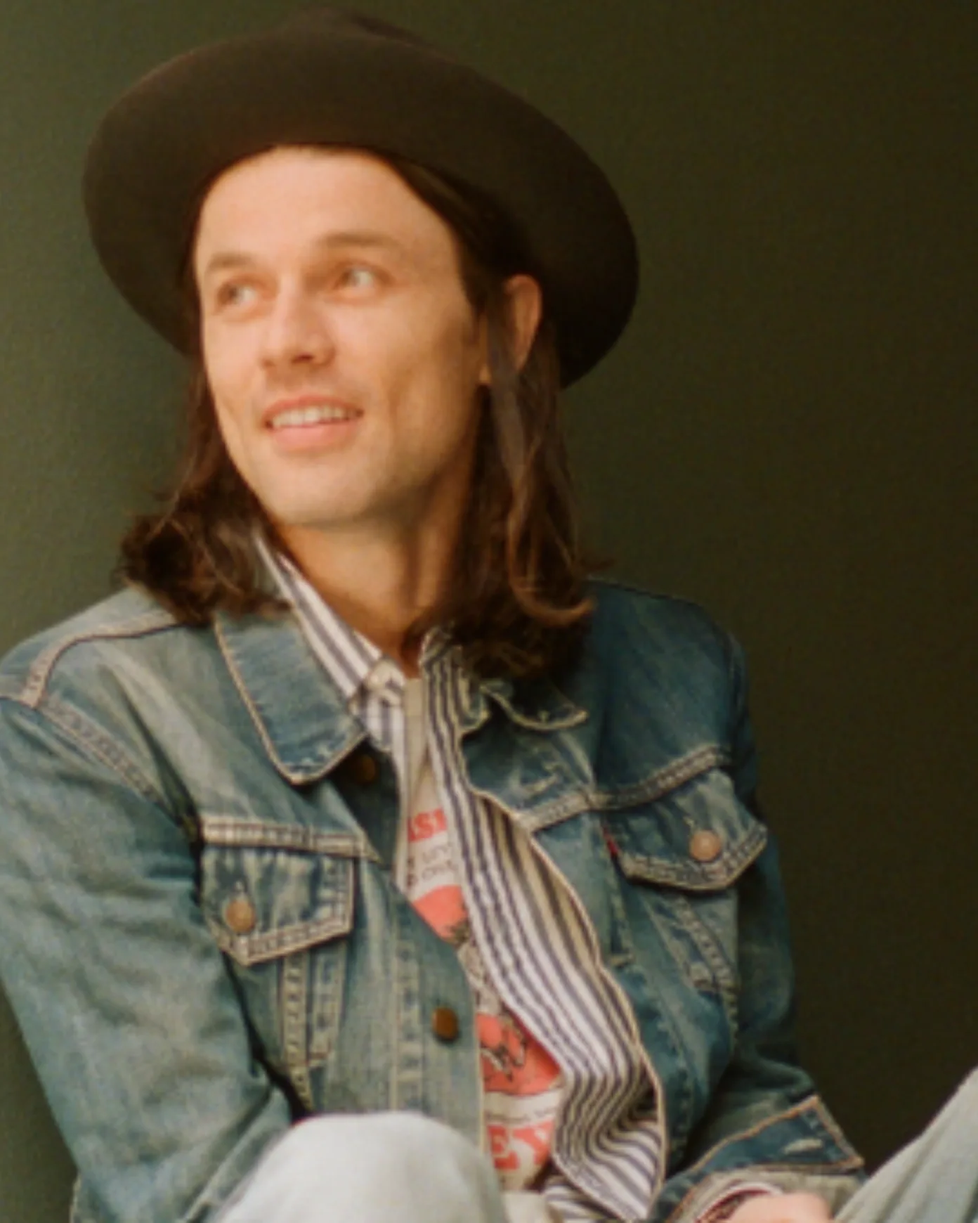 Smiling man with long hair and a brown hat looking away from the camera wearing a denim jacket with striped shirt beneath it sitting in front of a swamp green wall