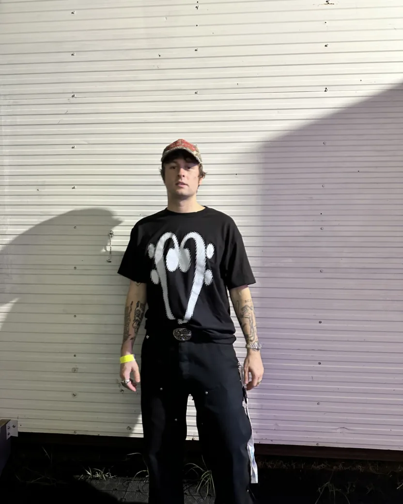 Tattooed man wearing a baseball cap and black clothes standing in front of a white textured wall