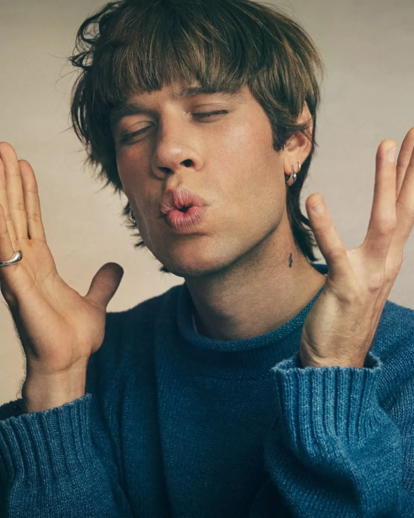 Man in blue knitted sweater holding his hands up next to his face, his eyes are closed and his lips are pouched