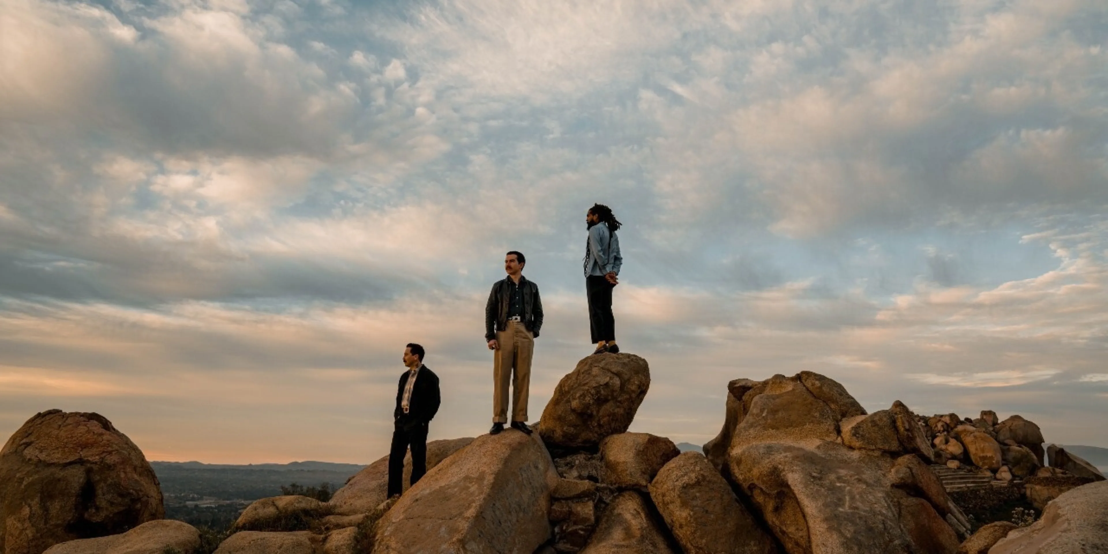 A picture of three man standing on rocks in front of a sky full of clouds during golden hour.