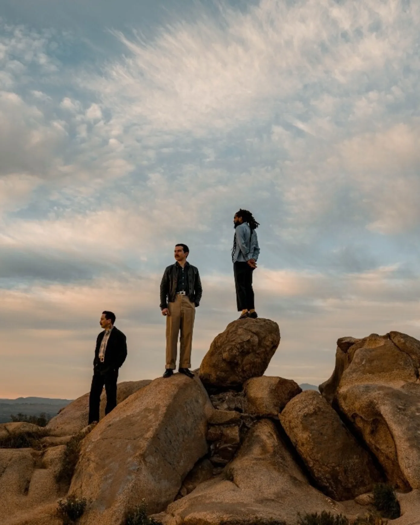 A picture of three man standing on rocks in front of a sky full of clouds during golden hour.